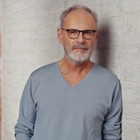 Ein Mann mit Brille und Pullover lehnt an einer Wand und schaut in die Kamera