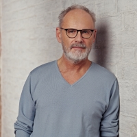 Ein Mann mit Brille und Pullover lehnt an einer Wand und schaut in die Kamera