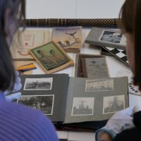 Zwei Personen sitzen an einem Tisch vor verschiedenen historischen Materialien wie Büchern und Orden und blicken in ein Fotoalbum.