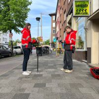 Zwei junge Menschen stehen mit Messinstrumenten an einer Straße an Stolpersteinen.