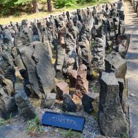 Auf einer freien Fläche im Wald befindet sich ein Denkmal: Steine verschiedener Höhen sind in einem Rechteck aufgestellt, im Vordergrund liegt ein Stein mit dem Schriftzug 'Münster' auf dem Boden.