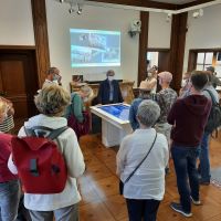 Ingrid Lueb in der Ausstellung an einem Touchtisch, von dem aus historische Fotos der Personen, die in der Villa gelebt und gearbeitet haben, an die Wand projiziert werden. Um sie herum stehen Teilnehmende der Führung.