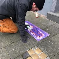 Ein junger Mann sprüht mit Kreide die Aufschrift #DeportationenSichtbarMachen neben Stolpersteine