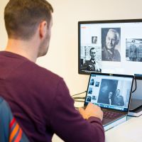 Ein Mann sitzt an einem Schreibtisch vor zwei Computer-Bildschirmen. Auf diesen sind historische Fotografien von Personen geöffnet.
