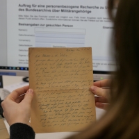 Eine Frau sitzt an einem Computerbildschirm. Auf dem Bildschirm ist ein Rechercheformular eines Archivs geöffnet. Die Frau hält einen handgeschriebenen Brief in der Hand.