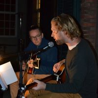 zwei junge Männer sitzen singend mit Gitarren auf der Veranda der Villa