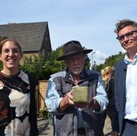 drei Menschen stehen nebeneinander, der Künstler in der Mitte hält einen Stolperstein in die Kamera