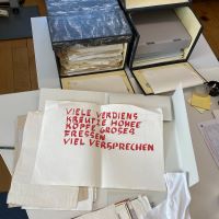 Auf einem Schreibtisch augebreiteter Inhalt eines Archivkartons. Im Zentrum steht ein Gedicht mit roter Aufschrift: "Viele Verdiens
kreutze hohee
Köpfe groses
Fressen
Viel Versprechen"