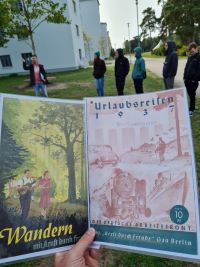 zwei historische Plakate der Organisation "Kraft durch Freude" werden in die Kamera gehalten, im Hintergrund Teilnehmende der Führung vor dem historischen Gebäude