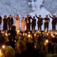 Menschen stehen auf einer Bühne vor einer Leinwand mit einem historischen Foto eines Kindes. Sie halten Lichter in die Höhe, ebenso wie das Publikum