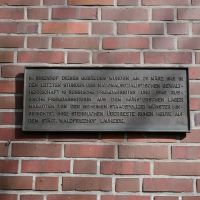Ein Schild mit Text hängt an einer Mauer.
Es erinnert an Verbrechen.
Und an die Toten.