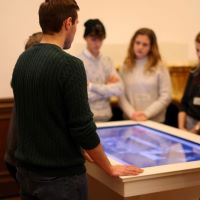 Menschen stehen in der Villa ten Hompel.
Sie stehen um einen Bild-Schirm.
Ein Mann erklärt etwas.
Die anderen hören zu.