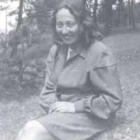 Eine Frau sitzt auf einer Wiese.
Hinter ihr stehen Bäume.
Sie lächelt.