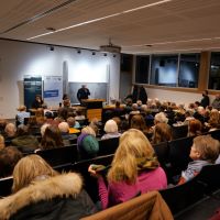 Das Publikum der Lesung sitzt in einem Hörsaal. Die Plätze sind voll belegt. Vorne an der Tafel sitzen Jennifer Teege und Oliver von Wrochem, während Thomas Köhler gerade spricht.