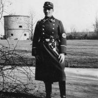 In dem Schwarzweißfoto sieht man einen Mann in einem Park. Er trägt eine dunkle, knielange Uniform der SA, an seiner Hüfte hängt ein Dolch. Er blickt direkt in die Kamera, die Hälfte seines Gesichts liegt wegen der Mütze auf dem Kopf in einem starken Schlagschatten.