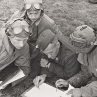 Vier Männer in Uniform liegen auf dem Boden um ein Blatt Papier herum, auf das zwei etwas einzuzeichnen scheinen, während die anderen beiden zusehen.