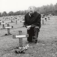 Ein Mann in einem Gräberfeld auf einem Soldatenfriedhof kniet vor einem Grab, die Hände gefaltet, den Kopf nach vorne gesenkt. Neben dem Grabkreuz ist ein Blumenstrauß niedergelegt worden.