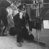 Ein SS-Mann in Uniform kniet an einem Zaun, wo er ein Feldtelefon aufgehängt hat, dessen Hörer er gerade ans Ohr hält.