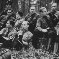 Zehn Männer in SS-Uniform sitzen gemütlich auf einem Waldboden, die meisten haben die Kopfbedeckungen abgenommen. Einer hat eine Zigarette und einen Flachmann in der Hand.