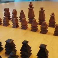 Auf einem Holztisch steht ein vollständiges Set an Schachfiguren aus Holz, die einen heller und die anderen dunkler lackiert. Die Figuren sind stilisiert und mit groben Kanten angedeutet.