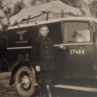 Ein etwas unscharfes Schwarzweißfoto von einem Mann in dunkler Uniform, einem schmalen Schnurrbart direkt unter der Nase. Er lehnt an einem großen Auto, auf dem neben einer Ziffer auch das Logo der Deutschen Reichspost angebracht ist.