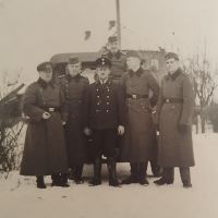 Auf dem Gruppenfoto sind sechs Männer zu sehen: Johann in der Mitte in dunkler Uniform und Kopfbedeckung steht frontal zur Kamera. Um ihn herum stehen fünf weitere Männer in Uniform, die Mäntel reichen bis zu den Knöcheln, vier von ihnen tragen Schiffchen, der letzte eine ähnliche Kopfbedeckung wie Johann. Sie stehen auf einer Straße mit Schnee, im Hintergrund sind ein Auto und Wohnhäuser zu sehen.