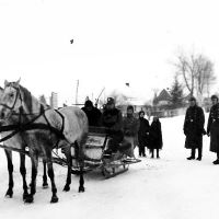 Auf einer verschneiten Straße steht ein großer Schlitten mit zwei Pferden, in denen Menschen in Uniformen sitzen. Rechts daneben stehen weitere vier Männer in Uniform und mit Kopfbedeckungen, im Hintergrund sind zwei Kinder sowie eine Wohnsiedlung zu sehen.