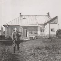 Ein Mann in Uniform steht vor einem einstöckigen Wohnhaus auf einer Wiese und blickt in die Kamera. Die rechte Hand hat er in die Hosentasche gesteckt.