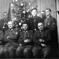 Zwölf Polizisten in Uniform stehen in einem Wohnraum für ein Gruppenfoto zusammen, die vordere der beiden Reihen sitzt. Einige Männer haben die Arme verschränkt, die meisten blicken neutral bis streng in die Kamera. Im Hintergrund steht ein geschmückter Weihnachtsbaum.