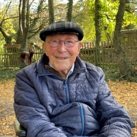 Ein älterer Herr in Daunenjacke und mit Schiebermütze und Brille blickt lächelnd in die Kamera. Im Hintergrund ist ein herbstlicher Garten zu sehen.