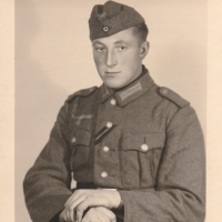 Ein junger Mann in Wehrmachtsuniform sitzt für ein Porträt. Er blickt direkt in die Kamera, die Hände hat er übereinandergeschlagen.
