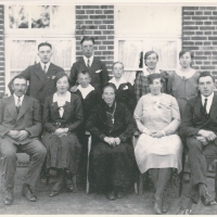 Elf Personen sitzen und stehen für ein Gruppenfoto vor einem Wohnhaus. Sie tragen Anzüge und Kleider, die meisten blicken ernst in die Kamera.