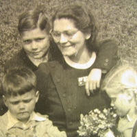 Auf dem Gruppenfoto sieht man eine lächelnde Frau mit Brille, die zur Seite auf eines ihrer drei Kinder blickt, die um sie herum sitzen. Ein Junge hinter ihr hat den Arm um sie gelegt, das jüngere Mädchen mit hellem Jahr blickt auf einen Blumenstrauß, den sie in Händen hält, und das dritte Kind, ein Junge, blickt mit gerunzelter Stirn nach unten.