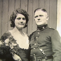 Ein junger Mann und eine junge Frau stehen für ein Hochzeitsfoto nebeneinander. Er trägt Uniform, steht gerade da und blickt über die Kamera hinaus; sie trägt ein dunkles Kleid mit hellem Kragen, hat einen Blumenstrauß im Arm und blickt direkt in die Kamera.