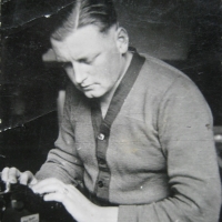 Ein junger Mann mit akkurat gescheiteltem Haar sitzt vor einer Schreibmaschine und blick darauf, die Stirn nachdenklich gerunzelt.