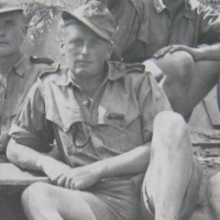 Ein junger Mann in Uniform mit kurzen Hosen sitzt lässig auf dem Boden, ein Bein angewinkelt, die Hände verschränkt, und blickt in die Kamera.