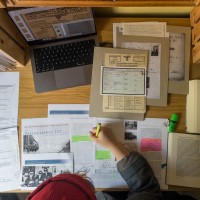 An einem Schreibtisch sitzt eine Person über Ausdrucke und Bücher zur NS-Zeit gebeugt.