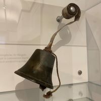 Eine ungefähr faustgroße Glocke hängt in einer Vitrine. Das Metall ist schon matt geworden, ein Stück Kordel ist über dem Glockenkörper befestigt, um sie anschlagen zu können.