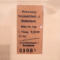 Eine Fahrkarte in einer Vitrine aus einfachem braunem Papier. Darauf steht: 'Personenzug Theresienstadt-Bausch A - Bodenbach. Gültig vier Tage. 3. Klasse 2,20 RM, 53 km.'