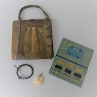 Auf einer weißen Oberfläche liegt eine kompakte Handtasche mit kurzem Henkel, Schmuckstücke und ein Nehset aus blaugelbem Karton.