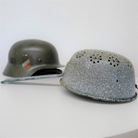 Das Küchensieb liegt im Vordergrund, ein Stahlhelm in einem dunklen Grünbraun im Hintergrund. Auf ihm ist ein schwarz-weiß-rotes Wappen zu sehen.