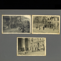 Auf einer Fotoalbumseite sind drei Fotos eingeklebt. Auf dem ersten ist eine Häuserzeile zu seehn, in der ein Haus vollständig eingestürzt ist. Auf der Straße davor steht eine Frau vor einem Schuttfeld. Auf dem zweiten Foto blickt man auf eine weitere Straße mit Häuserzeile: Eines der Häuser brennt, die anderen sind verkohlt, davor sind Menschen unterwegs; zwei von ihnen tragen einen Sarg. Auf dem dritten Foto blickt man auf eine Straße, auf der Menschen unterwegs sind. Zwei Tierkadaver, fast vo