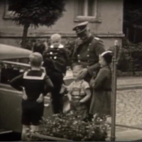 Auf einer etwas unscharfen Filmaufnahme sieht man mehrere Personen auf einer Straße neben einem Auto mit offener Tür. Drei Kinder stehen um ihn herum, eine Frau hält ein Kleinkind im Arm.