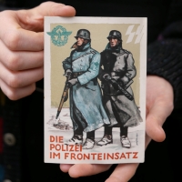 Auf einer Postkarte ist eine Malerei abgebildet mit zwei Polizisten in Uniform, mit Helmen und Waffen in der Hand in einer verschneiten Landschaft. Darunter steht 'Die Polizei im Fronteinsatz'.