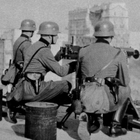 Drei Männer in Uniform sitzen auf einem Flachdach hinter einem Maschinengewehr und blicken in die Ferne. Im Hintergrund sind zerstörte Gebäude zu erkennen.