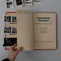 Ein Buch liegt aufgeschlagen auf einem Tisch und jemand blättert darin. Sichtbar ist das Schmutzblatt mit dem Titel des Buchs sowie Fächer im Buchdeckel, aus denen Fotos und die Brille hervorschauen.