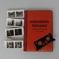 Auf dem Buch mit rotem Einband liegt der zusammengefaltete Stereobetrachter, links daneben vier Stapel der Stereofotos, auf denen Landschaften, Stadtansichten, Festbeleuchtung und eine sprechende Person zu sehen sind.