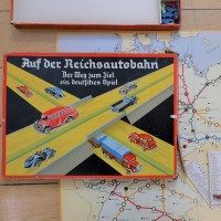 Auf einem aufgeklappten Spielfeld mit einer Landkarte Deutschlands liegt die Verpackung des Spiels. Darauf fahren vor schwarzem Hintergrund bunte Autos auf gelben Autobahnen.
