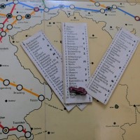 Auf dem Spielbrett liegen drei Spielkarten und eine Figur in Form eines Autos. Auf den Karten stehen verschiedene Orte des damaligen Deutschlands.