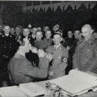 Auf einem Schwarzweißfoto steht eine große Gruppe von Männern in unterschiedlichen Uniformen in einem Festsaal. Einige lächeln. Im Vordergrund steht Joseph Goebbels und hebt einen Pokal zum Mund, neben ihm ist ein Tisch mit einem großen aufgeschlagenen Buch zu erkennen.